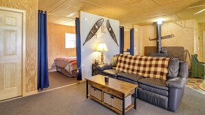 Un salón acogedor con un sofá de cuero, una mesa de centro de madera y una lámpara. Se ve una cama con una colcha a cuadros al fondo.