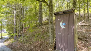Un baño exterior de madera con un adhesivo de luna creciente, rodeado de un área boscosa con un sendero de grava.
