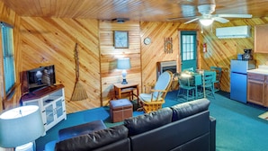 Interior acogedor de una cabaña con detalles de madera, ventilador de techo, televisión, chimenea, comedor y cocina.
