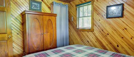 Um quarto com painéis de madeira, uma cama, um guarda-roupa de madeira, uma janela com persianas e duas molduras com quadros na parede.