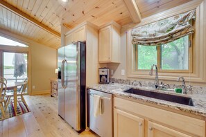 A kitchen with wooden cabinets, a stainless steel refrigerator, a dishwasher, a granite countertop, and a window with a curtain.