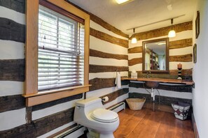 Een rustieke badkamer met een houten wand van boomstammen, een raam met jaloezieën, een toilet, een wastafel met spiegel erboven en een hanglamp.