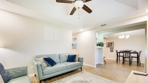 Ein Wohnzimmer mit einem hellblauen Sofa, einem gemusterten Teppich, Holzböden, einem Deckenventilator und einem Essbereich mit Tisch und Stühlen.