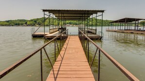 Un muelle de madera que lleva a una zona cubierta con asientos, con vista a un lago tranquilo y una ladera boscosa al fondo.