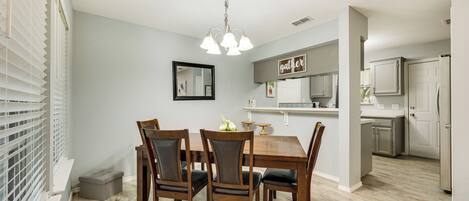 A dining area with a wooden table and chairs, a chandelier, and a kitchen in the background.
