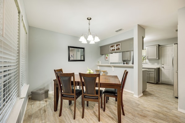A dining area with a wooden table and chairs, a chandelier, and a kitchen in the background.