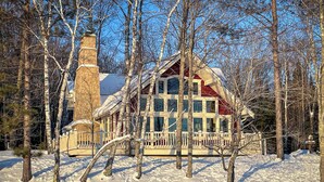 Una casa grande de varios pisos con una chimenea destacada, rodeada de terreno y árboles cubiertos de nieve.