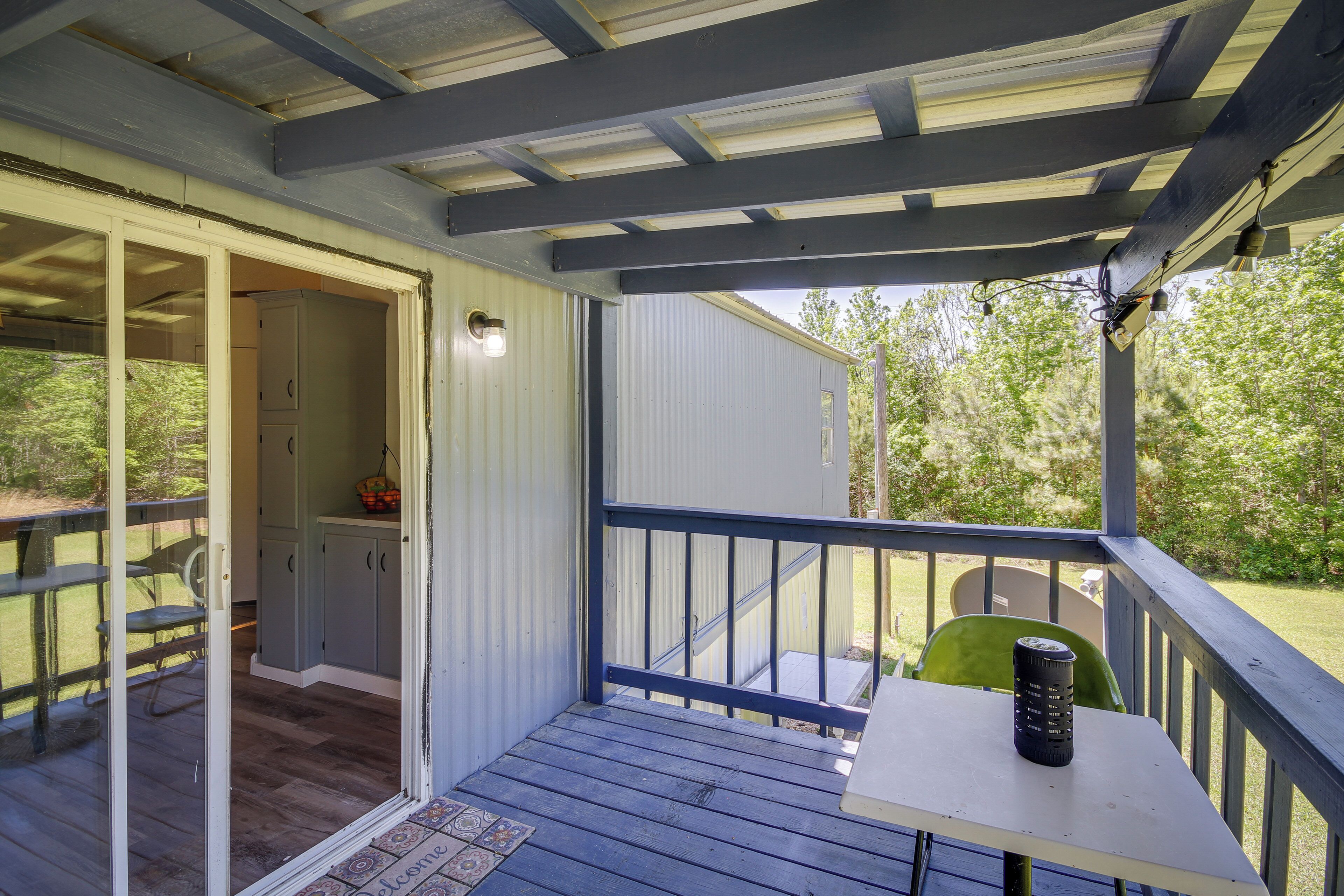 A covered outdoor area with a table and chairs, a door leading to an interior space, and a view of greenery outside.