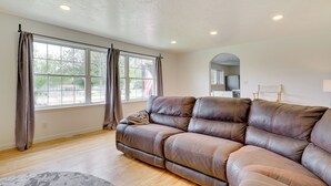 Un salon avec un canapé en cuir marron, de grandes fenêtres avec des rideaux, un plancher en bois et un petit tapis.