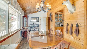 A wooden interior with a dining table, chairs, a chandelier, a mounted deer head, and large windows.