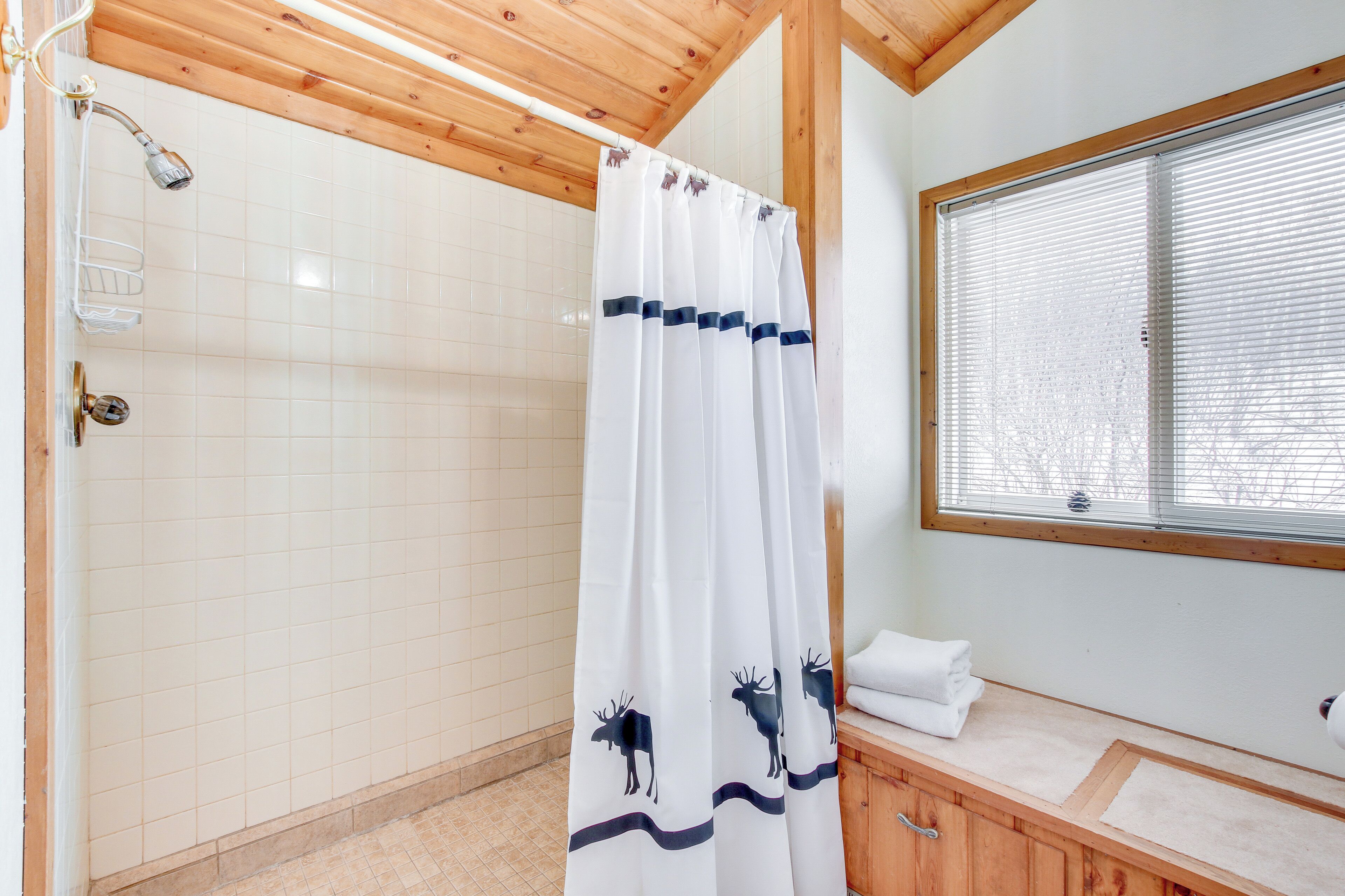 A bathroom with a shower, a window with blinds, a wooden ceiling, and a curtain with a moose design.