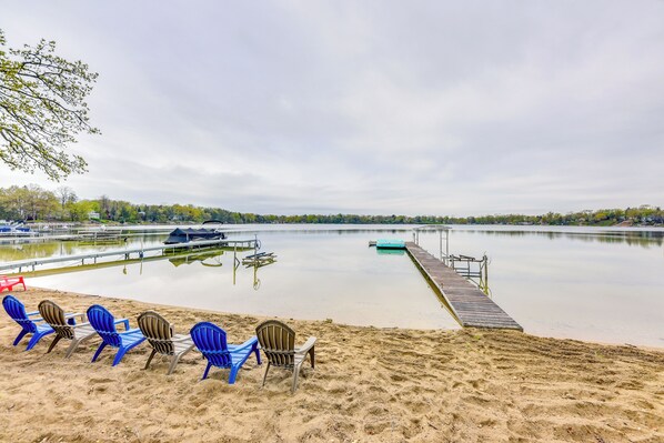 A lakeside beach with blue and brown chairs, a wooden dock, and a calm water body. - Lakefront Burlington Vacation Rental: Dock + Beach (Burlington)