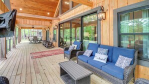 A covered outdoor seating area with wicker furniture, a red rug, and a view of the forest. - Gorgeous Drasco Home Near Greers Ferry Lake! (Drasco)