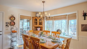 A dining area with a wooden table set for six, surrounded by wooden chairs. A chandelier hangs from the ceiling, and there are two windows with blinds. - Near Highlands: 36-acre Scaly Mountain Lodge (Scaly Mountain)