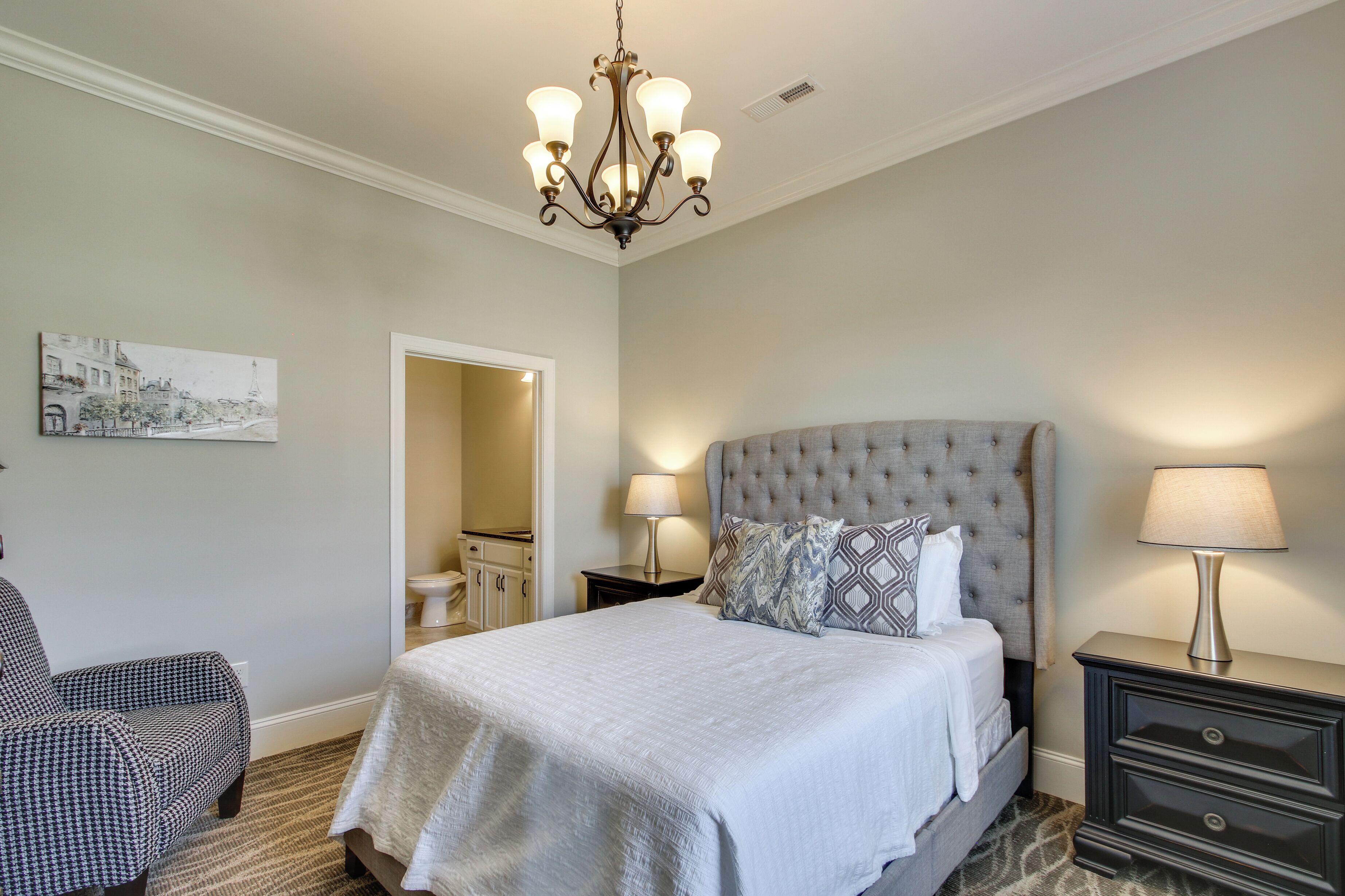A bedroom with a large bed, a chandelier, a nightstand, a chair, and a painting on the wall.