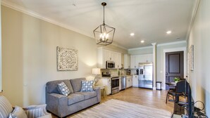 Una sala de estar moderna con un sofá gris, una zona de comedor con mesa y sillas, y una cocina con armarios blancos y electrodomésticos de acero inoxidable.