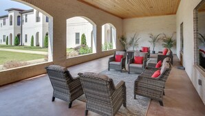 Un patio techado con mobiliario de mimbre y cojines rojos, ventanas amplias y vistas a un edificio y césped.