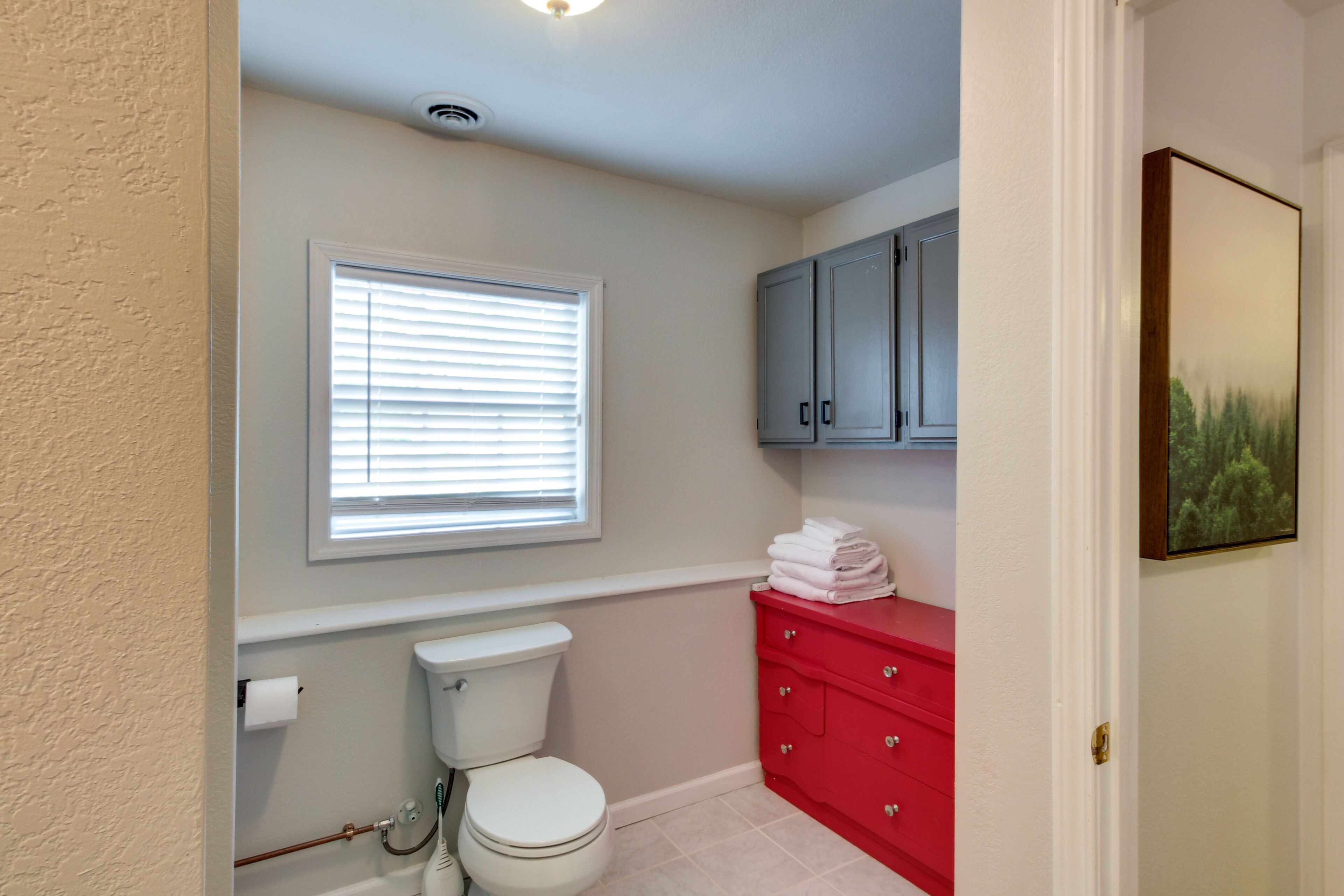 A bathroom with a toilet, a window with blinds, a red vanity with drawers, and a cabinet above the vanity.