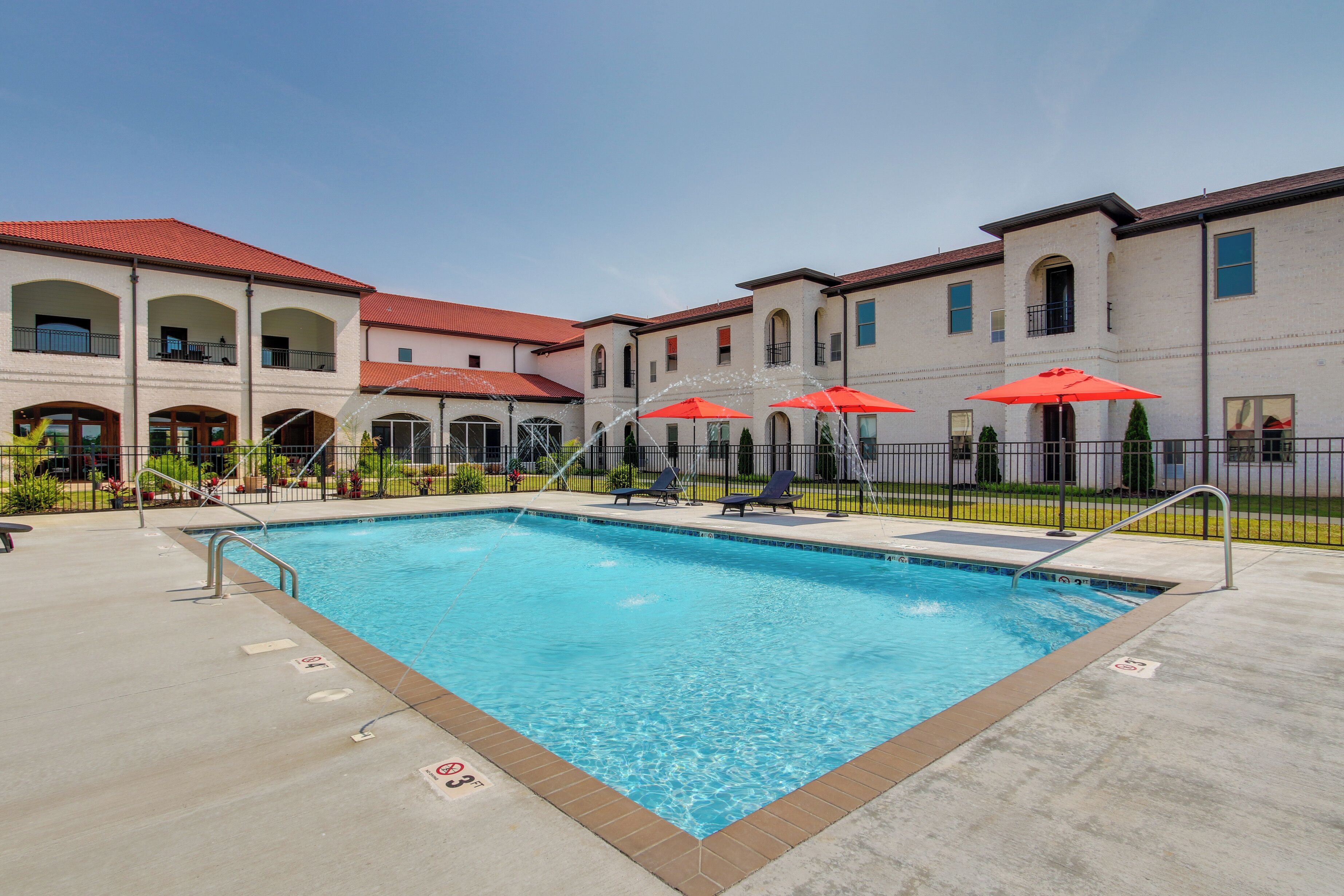 A swimming pool with a clear blue water, surrounded by a concrete deck and a fence. There are red umbrellas and lounge chairs around the pool.
