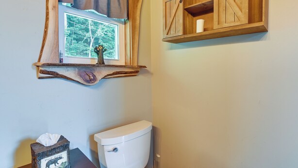 A bathroom with a rustic wooden shelf above a window, a white toilet, and a tissue box labeled "Post Card Bear Crest."