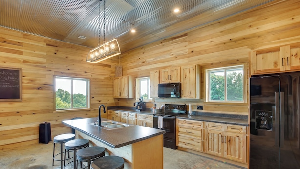 A modern kitchen with wooden cabinetry, a central island with stools, stainless steel appliances, and large windows. - Burkesville Cabin, 3 Mi to Dale Hollow Lake! (Burkesville)