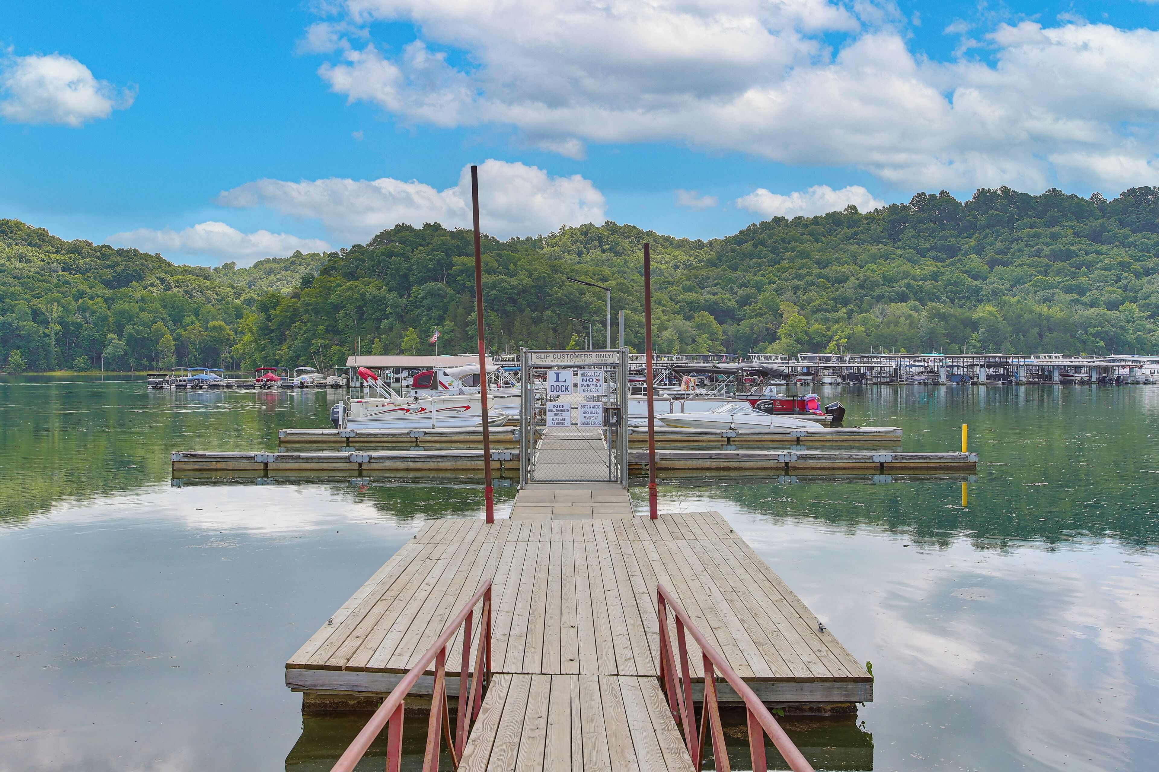 A wooden dock leading to a marina with boats, a dock sign, and a forested hill in the background.