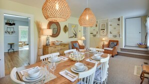 A dining area with a wooden table set for four, surrounded by white chairs. Two large woven pendant lights hang above the table. - Mobjack Bay Rental on Private Cove! (Bohannon)