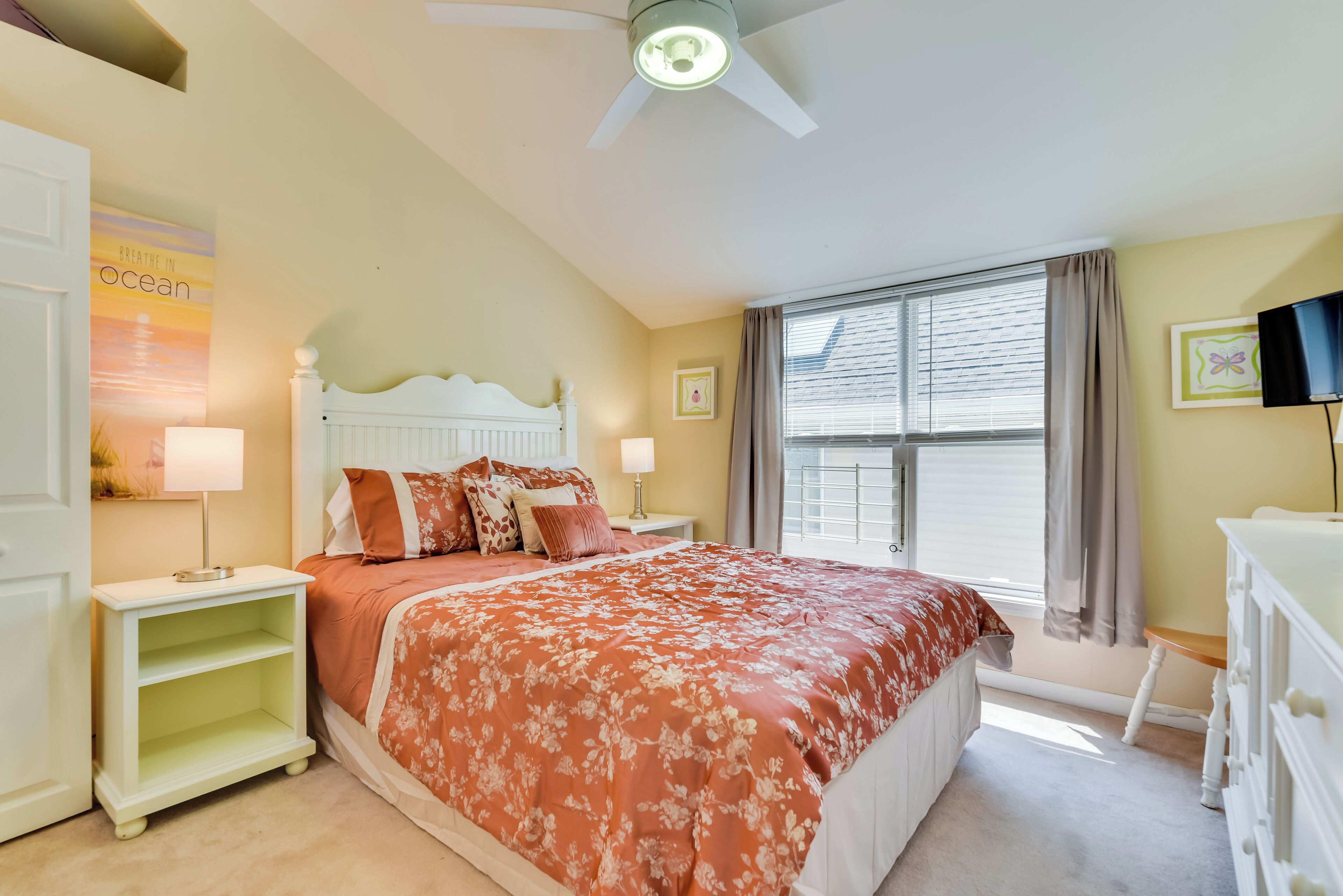 A bedroom with a bed, nightstands, a ceiling fan, a window with blinds, and a wall-mounted TV.
