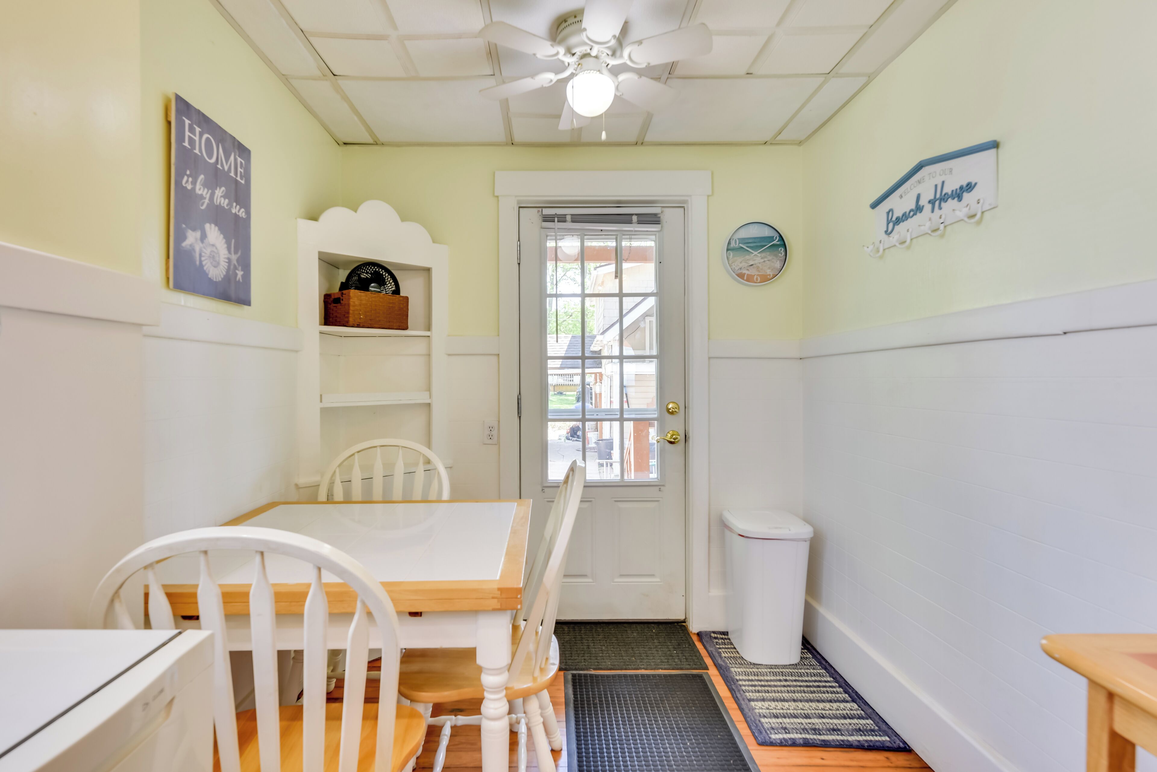 A dining area with a white table, chairs, and a ceiling fan. There is a door with a window, a trash can, and a rug on the floor.
