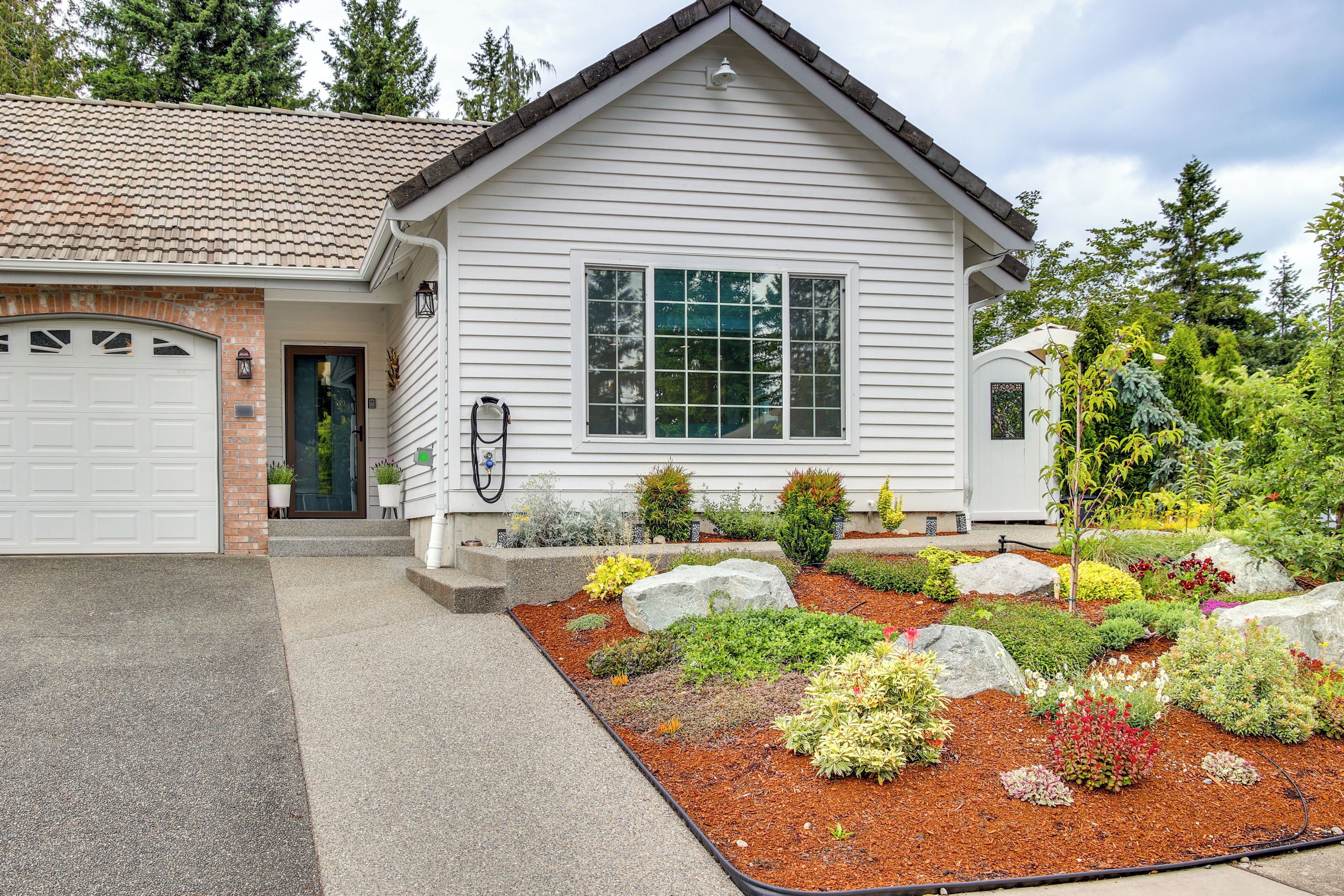 A single-story house with a garage, a front porch with potted plants, and a landscaped garden with various shrubs and a rock centerpiece.