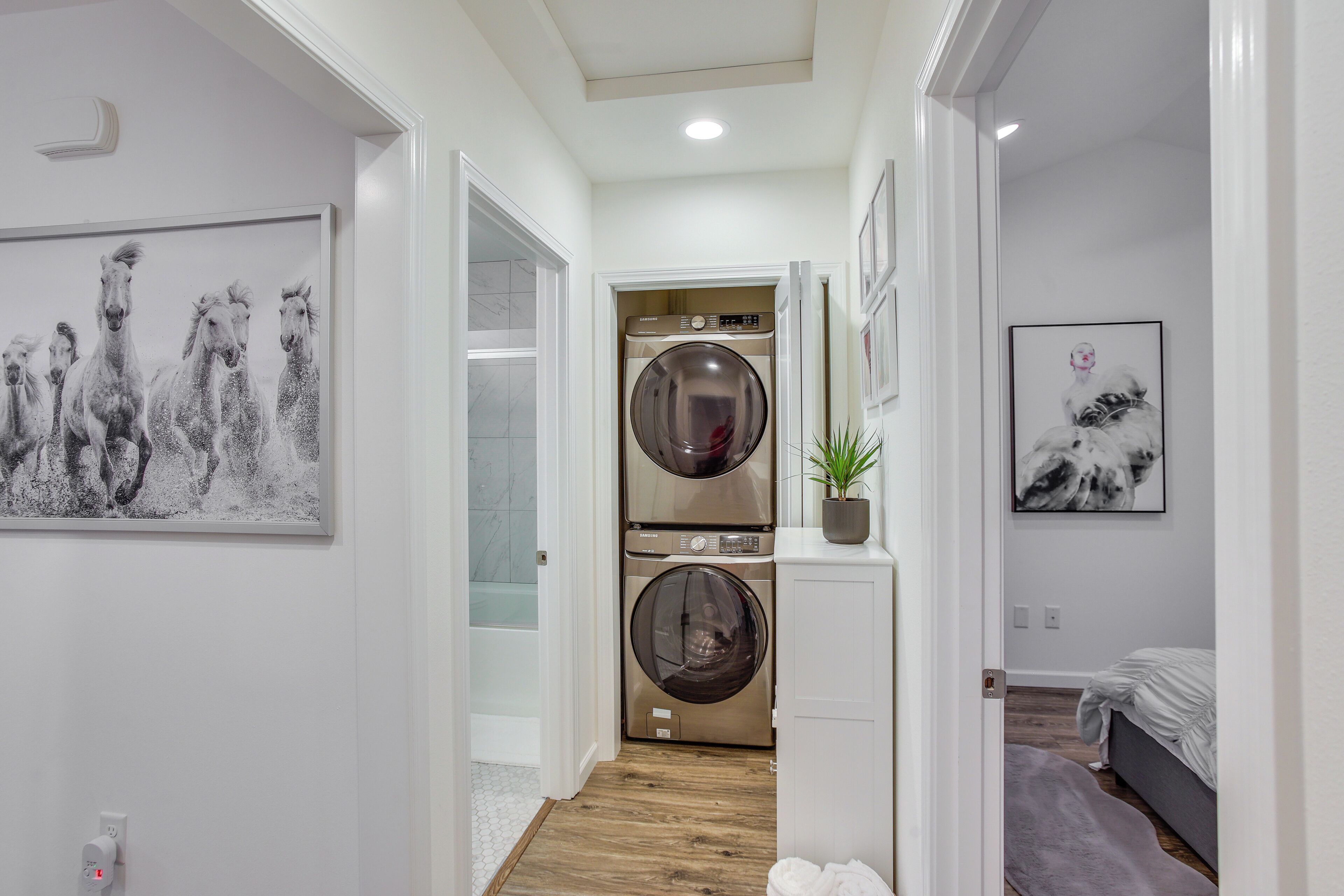 A laundry room with two stacked front-loading washers and dryers, a potted plant, and artwork on the walls.