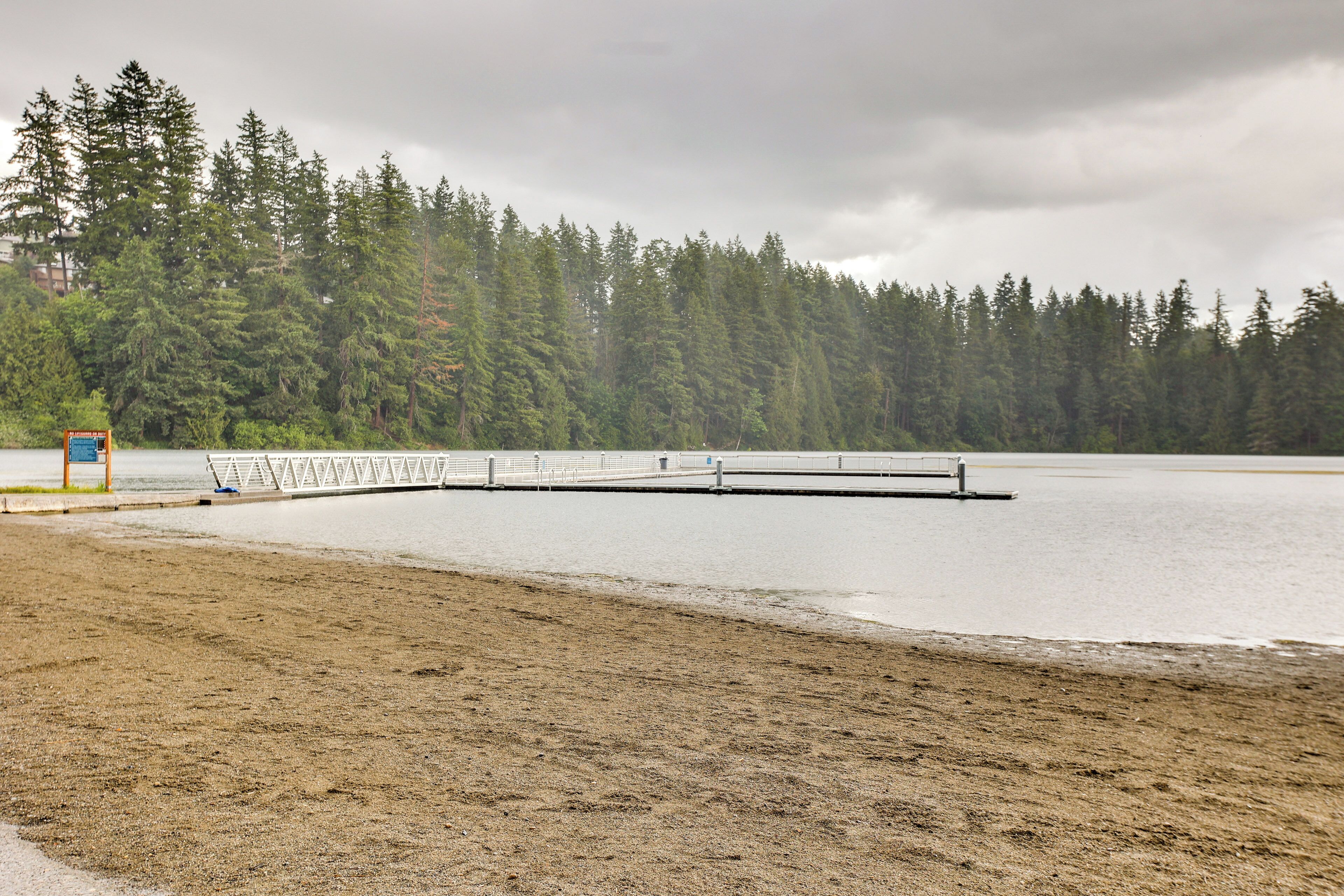 A sandy beach with a wooden pier extending into a calm lake, surrounded by dense forest.