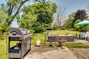 A backyard with a barbecue grill, a grill cart, a table with outdoor seating, and a large planter with flowers. - Walworth Home w/ Hot Tub - Near Golf Courses! (Walworth)
