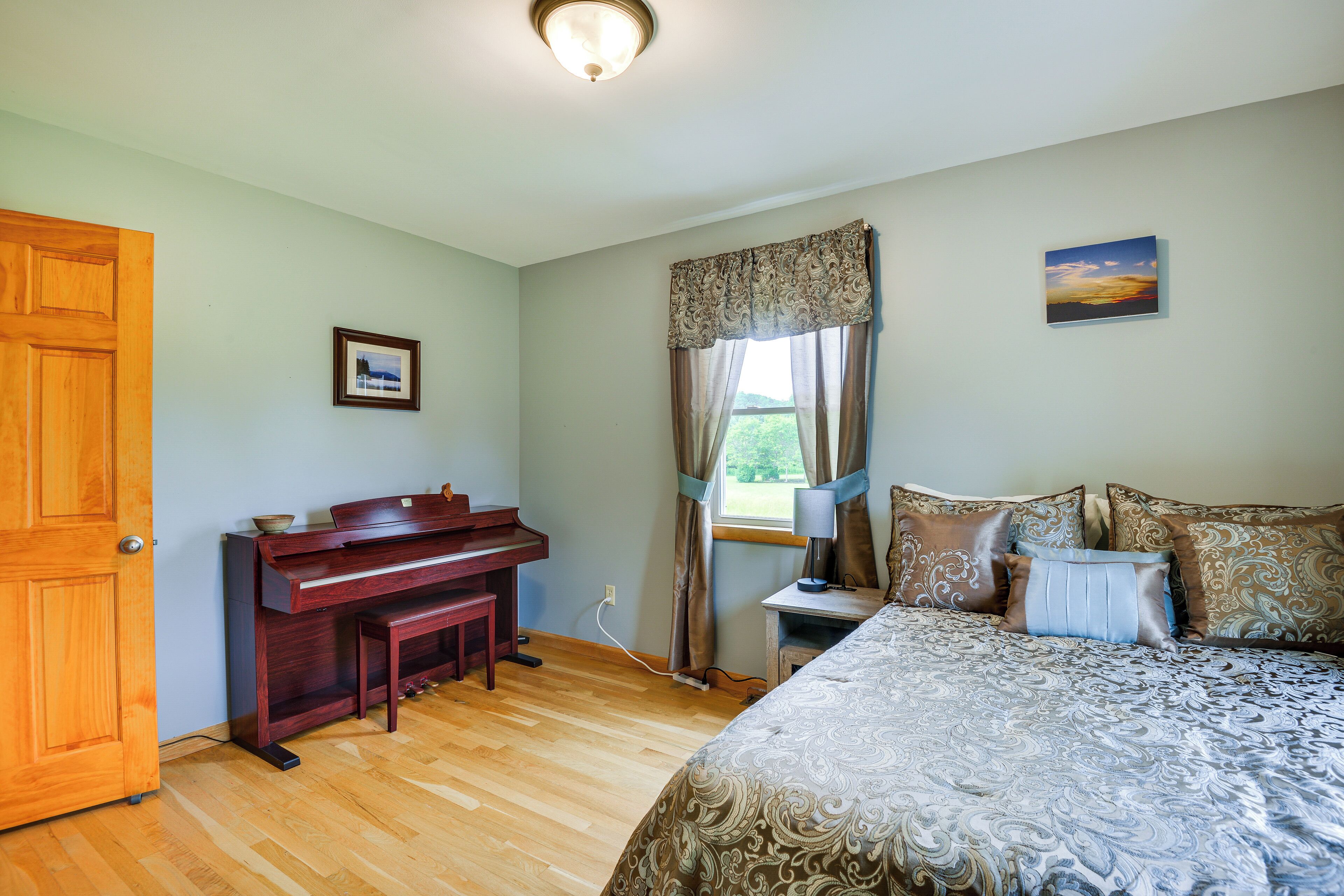 A bedroom with a wooden door, a piano, a bed with patterned bedding, a nightstand with a lamp, and a window with curtains.