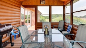 Ein Holzblockhaus-Interieur mit einem Glastisch, Stühlen, einem Grill und Blick ins Freie.