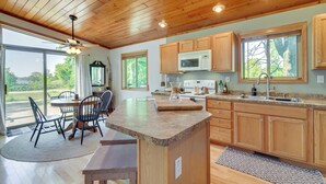 A spacious kitchen with wooden cabinets, a granite countertop, and a large island. There is a dining area with a table and chairs, and a view of the outdoors through large windows.