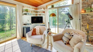 A cozy living room with a fireplace, a television, and a large window overlooking a forest.