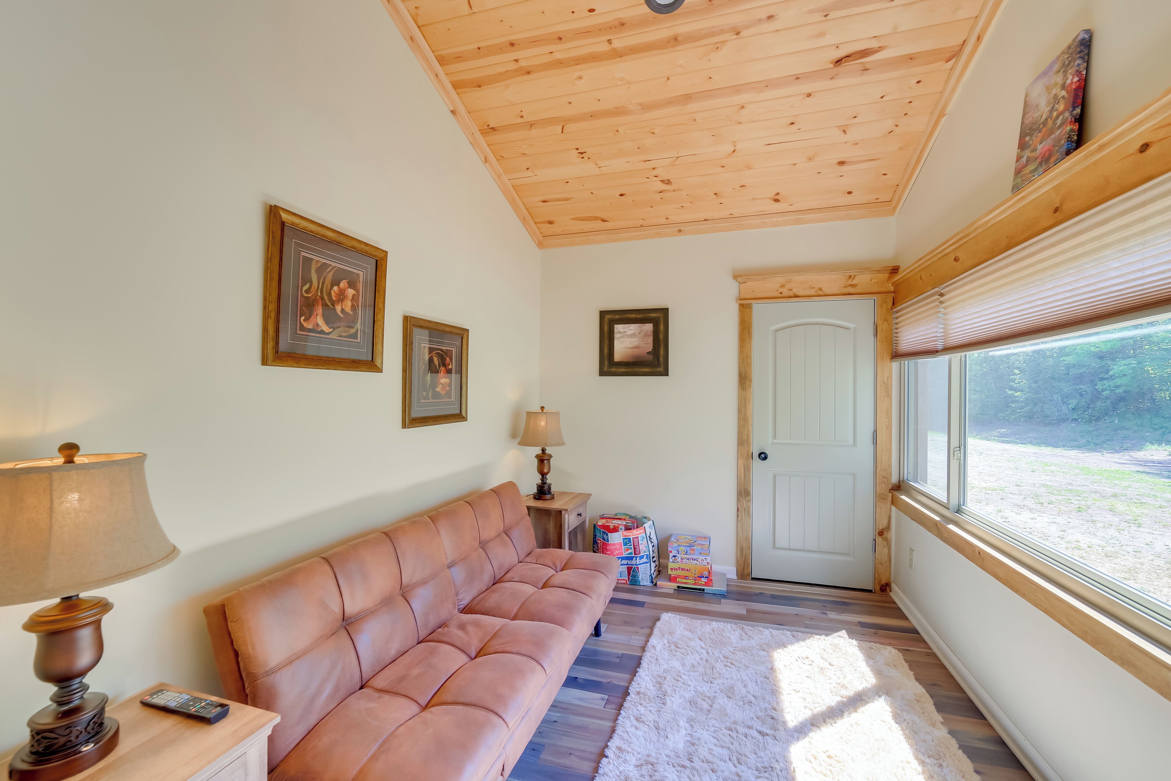 A cozy room with a wooden ceiling, a cushioned sofa, a lamp, framed pictures, a door, and a window with a view of greenery.