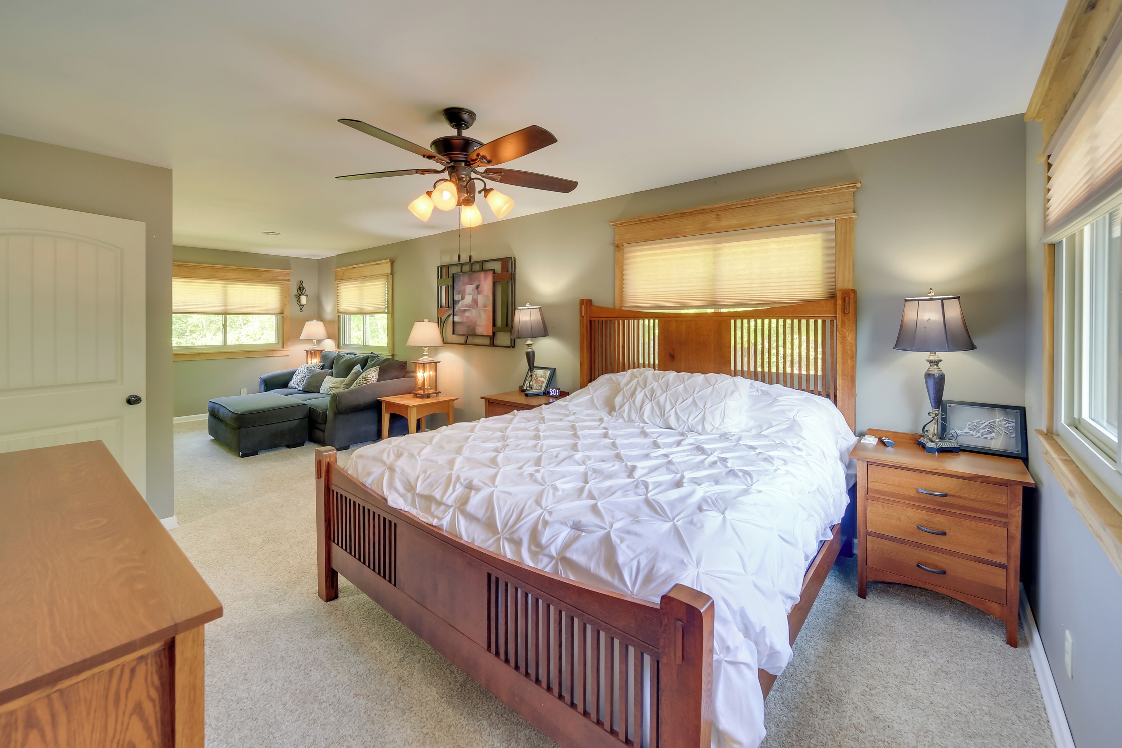 A bedroom with a large bed, a ceiling fan, two bedside tables with lamps, a wooden nightstand, and a window with blinds.