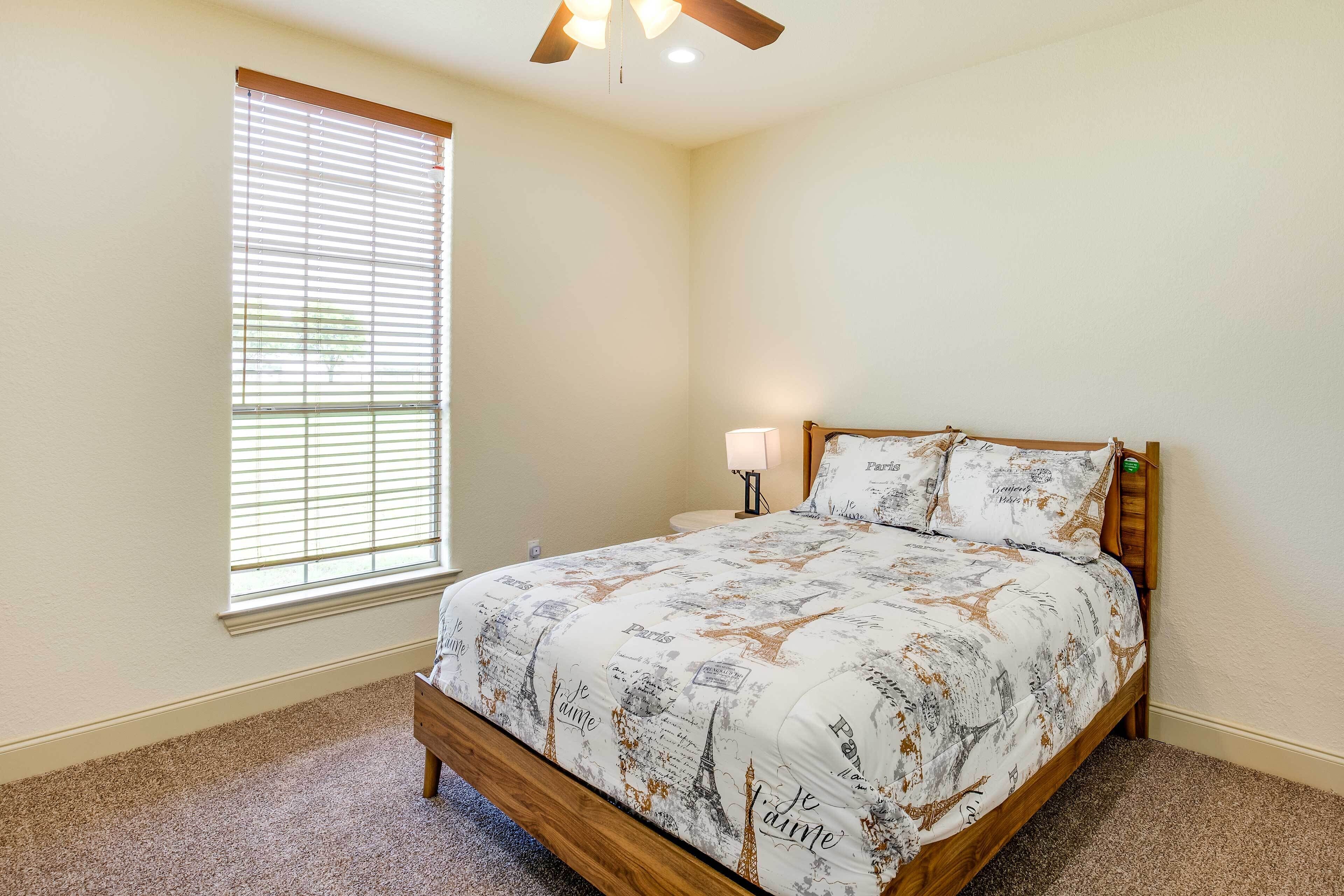 A bedroom with a bed, a window with blinds, a ceiling fan, and a nightstand with a lamp.
