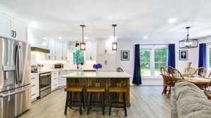 A modern kitchen with white cabinets, stainless steel appliances, wooden stools, and a dining area with a table and chairs. - Hot Tub Haven 2 Mi to Beach! Christy's Cottage (Snow Hill)