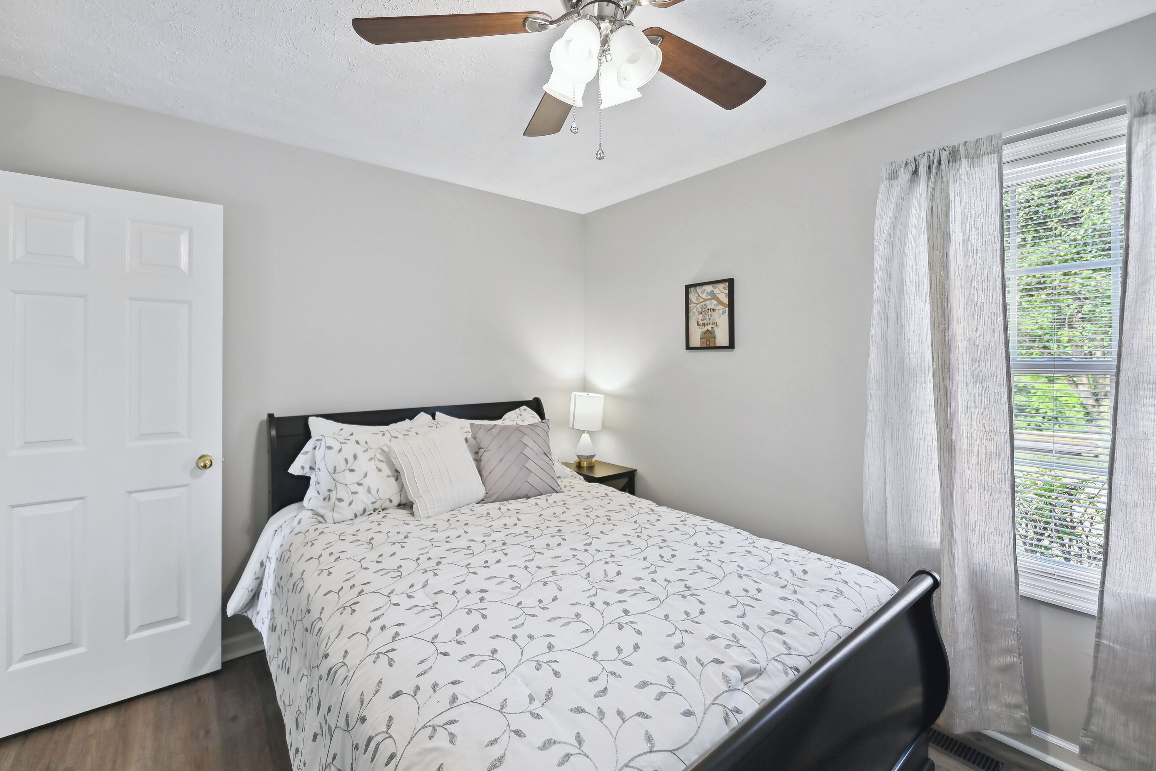 Une chambre à coucher comprenant un lit, un ventilateur de plafond, une table de chevet avec une lampe, une porte et une fenêtre avec des rideaux.