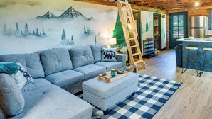 Un salon chaleureux avec un canapé gris, une table basse en bois et un tapis à carreaux. Un mur est décoré d’une fresque représentant un paysage de montagne.