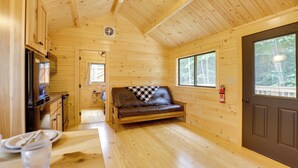 Interior de una cabaña de madera con sofá, zona de cocina y una puerta que da al exterior.