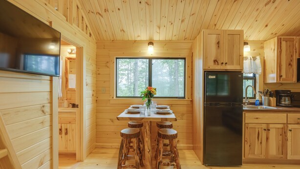 Interior de una cabaña de madera con una mesa de comedor, taburetes, un refrigerador y armarios de madera.