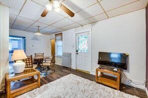 Ein Wohnzimmer mit Holzboden, einem weißen Teppich, einem Fernseher auf einem Holzbodenständer, einer Lampe auf einem Beistelltisch und einem Deckenventilator.