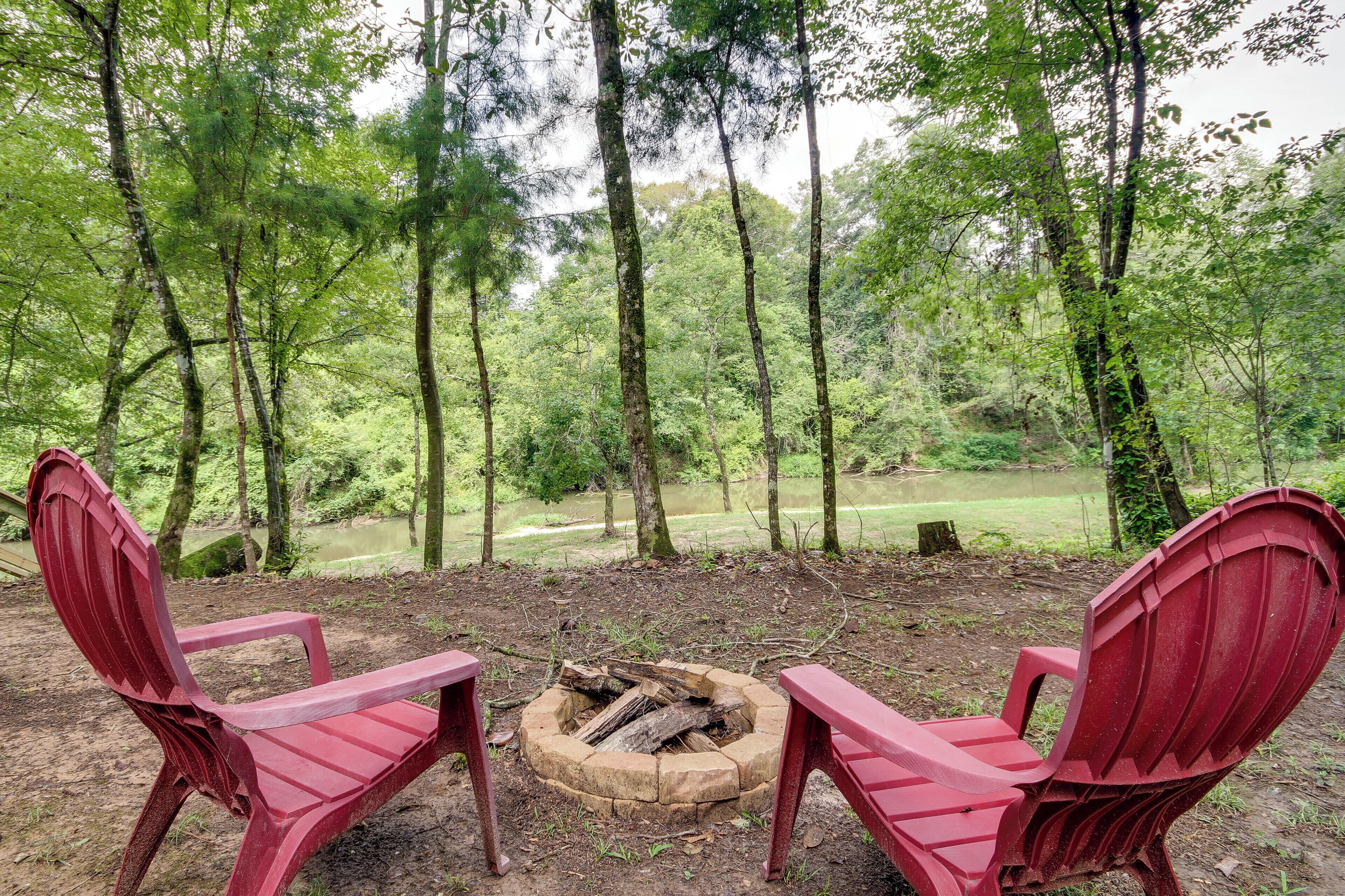 Two red Adirondack chairs facing a fire pit in a forested area with a view of a lake.