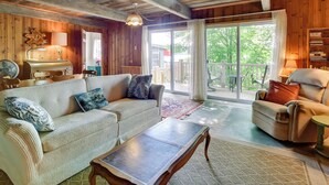 Un salon chaleureux avec un canapé, un fauteuil et une table basse. Un ventilateur et une lampe sont posés sur une table d’appoint. On peut voir un plafond en bois et une grande fenêtre donnant sur l’extérieur.