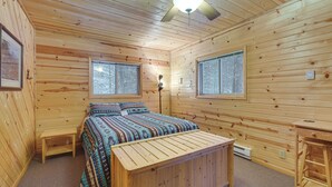 Ein Schlafzimmer einer Holzhütte mit einem Bett, zwei Fenstern, einem Deckenventilator und einem kleinen Tisch.