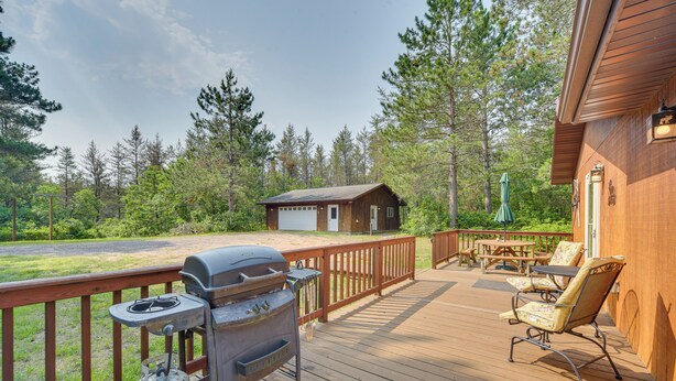 Eine Holzterrasse mit Grill, Sitzgelegenheiten im Freien und Blick auf eine Hütte in einem bewaldeten Gebiet.